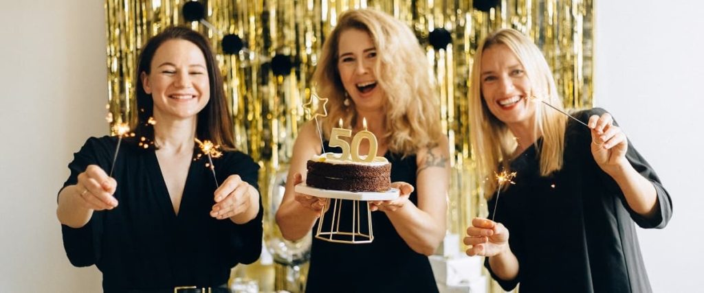 Femme fêtant son 50ème anniversaire, avec un gâteau dans les mains, tandis que ses amies tiennent des bougies à étincelles. 